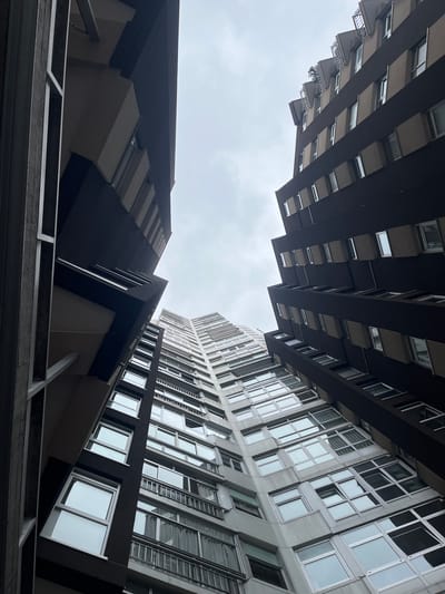 Looking upwards, the image captures the architecture of buildings in the city of La Coruña, Spain. Two buildings of varying architectural styles occupy most of the frame. The building on the left exhibits balconies and dark brown-colored construction, while the one on the right is a lighter gray with large windows, and what appears to be fire-escape ladders. The sky above is overcast, creating a muted light. The bottom edge of the frame includes a gray, metal framework.
