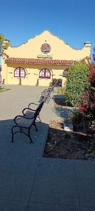 The image displays an exterior view of the historic Petaluma train depot, located in Petaluma, United States, under clear, bright daylight. The building features a yellow stucco facade with a distinctive scalloped parapet, a red tile roof, and two prominent arched windows with red frames and white grilles. The word "PETALUMA" is inscribed on the building's facade below a central emblem.
In the foreground, a row of at least four unoccupied public benches, constructed with black metal frames and wooden slats, are linearly arranged on a dark grey paved surface, facing the building. To the right of the paved area, a landscaped bed contains dark soil, green shrubs, and plants with reddish foliage. A small, light-colored concrete block is visible within this planter bed. The scene is devoid of individuals. Direct sunlight illuminates the scene, casting distinct shadows from the benches and the building, indicating a light source from the left of the frame.