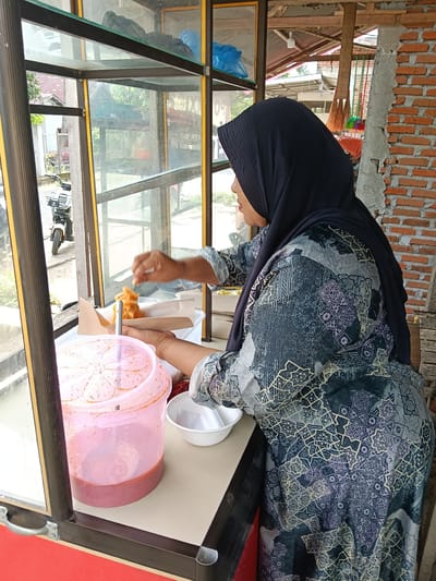 A woman wearing a dark hijab and a patterned dress is shown from the side, standing behind a glass display counter. She is holding a utensil and appears to be serving food from a paper container into a small white bowl. In front of her, on the counter, is a large pink plastic container filled with a reddish liquid, likely a sauce or drink. The display counter has a red base and a beige countertop. Through the glass, a motorcycle is visible outside. The background shows a brick wall and a wooden structure, suggesting an outdoor or semi-outdoor setting, possibly a street food stall. The overall scene depicts a moment of food service.