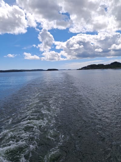 The image displays an expansive outdoor scene, likely from a moving boat, within the archipelagic environment of Mörkö, Sweden. The lower two-thirds of the vertical composition are occupied by a body of water, characterized by a prominent, turbulent white wake extending from the bottom center of the frame towards the distant horizon. The water on either side of the wake is dark blue-grey, with subtle surface ripples. Along the horizon, a series of low-lying, dark green, forested landmasses or islands are visible, stretching horizontally across the midground. The upper third of the image presents a bright blue sky filled with numerous white and grey cumulus clouds of varying sizes and shapes, indicating partial cloud cover with areas of direct sunlight. No individuals or specific man-made objects other than the implied vessel creating the wake are discernible. The lighting suggests a clear, bright daytime setting.