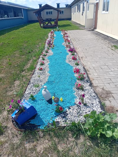 A decorative garden bed shaped like a river is featured, filled with bright blue gravel. Along its edges, white gravel and various colorful flowers, predominantly pink and purple petunias, are planted. In the foreground, a white stork figurine stands beside two small yellow chicks. To the left, a blue overturned planter with flowers spills out onto the blue gravel.
The garden bed is bordered by a dark material and is situated next to a paved walkway made of grey rectangular bricks. Beyond the walkway, a long, beige building with multiple windows and an open door is visible. To the left of the walkway, a well-maintained grassy area slopes upwards towards a dark blue building with large windows. In the background, a wooden gazebo with a table and benches can be seen on the grass.
The scene appears to be outdoors during the daytime under a bright, partly cloudy sky. The overall impression is one of a neatly arranged, decorative outdoor space, possibly part of a park or residential area. The location context is Пралетарскі сельскі Савет, Belarus. No text is clearly legible in the image.