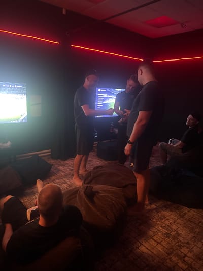In a dimly lit room with red neon lighting accents, four individuals are engaged in activities related to video gaming. Two men stand near a large screen displaying a sports game, one of whom appears to be holding a controller and interacting with the game. Another man is seated on a beanbag chair to the right, looking towards the screen. A fourth person is partially visible in the foreground, lying on a beanbag chair with a video game controller on their lap. The room has a modern, casual aesthetic, with beanbag chairs scattered on a patterned rug. The environment suggests a recreational space, possibly a gaming lounge or a private entertainment area. There is no identifiable information in the image that would place the scene in Beryozovka, Russia.