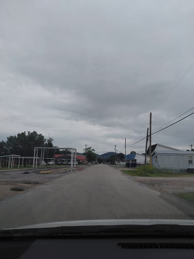 The image depicts a street scene in Stanton, United States of America, under an overcast sky. The road, which appears to be asphalt, shows signs of wear and tear, with several puddles reflecting the sky. On the left side of the road, there is a row of white structures, possibly carports or covered walkways, with some cars parked underneath. Beyond these structures, a large building with a red roof is visible, partially obscured by trees. Further down the street, more buildings and trees line the road, with a distant view of hills. On the right side of the road, a white mobile home with a blue roof is situated, accompanied by power lines running overhead. The scene is characterized by its muted colors and a general sense of quietness, suggesting it might be a residential or semi-commercial area.