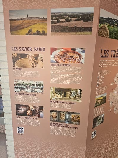 An angled informational display panel, light brown with a repeating white bean motif, is positioned in an indoor setting in Avignonet-Lauragais, France. The panel leans against a light-colored brick or stone column on its left side.
The display features multiple photographic prints and French text. The top section presents two landscape photographs: the left depicts a rural scene with green and golden fields leading to a distant town crowned by a prominent church steeple, identifiable as Avignonet-Lauragais; the right shows tilled fields and scattered trees under a hazy sky.
Below these, two main sections are titled "LES SAVOIR-FAIRE" (Know-how) on the left and "LES TRÉSORS" (Treasures) on the right (partially visible).
Under "LES SAVOIR-FAIRE," four distinct subjects are illustrated:
1. "LA POTERIE" (Pottery): A close-up of hands shaping clay on a potter's wheel, with finished ceramic pieces nearby.
2. "LES MARCHÉS DE PLEIN VENT" (Open-air Markets): A photograph of a vibrant outdoor market with numerous stalls and people.
3. "RÉVÉLATIONS VOYAGES D'ART" (Art Travel Revelations): An interior view of an art space featuring a large, intricate sculpture and other artistic elements. A black-and-white QR code is positioned below this description.
Under "LES TRÉSORS," three subjects are depicted:
1. "ZOOM SUR LE CASSOULET" (Zoom on Cassoulet): A close-up of a cassoulet dish in a brown ceramic bowl, garnished with herbs, on a table.
2. "LE CUIVRE HÉRIS EN LAURAGais" (Copper HERIS in Lauragais): An interior photograph displaying various copper items and a large decorative wall hanging, likely within a workshop or gallery.
3. "MUSÉE XXX ROBERT ET XX LA TAPISSERIE DU XXI SIÈCLE DU SUREZE" (Museum XXX Robert and XX Tapestry of the XXI Century from Sureze): An interior view of a museum exhibition space showcasing multiple large, colorful tapestries on the walls. A second black-and-white QR code is located below this section's text.
The overall environment suggests a well-lit exhibition space, possibly a museum or tourist information center, designed to inform visitors about the local culture, skills, and attractions of Avignonet-Lauragais and the Lauragais region. No individuals are directly interacting with the display within the frame.