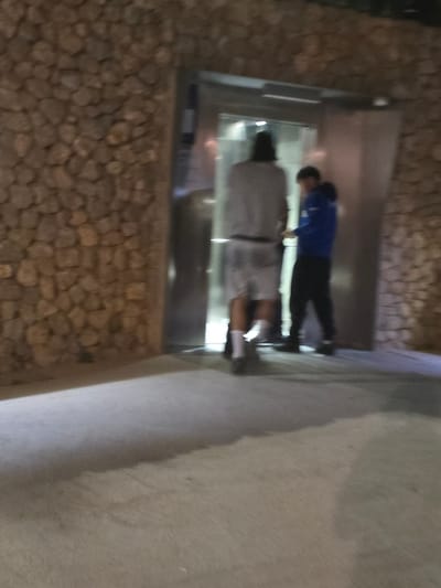 In Cornellà De Llobregat, Spain, two individuals stand near an elevator. The person on the left, facing away from the camera, is wearing a light-colored long-sleeved shirt and shorts, with white socks and dark shoes. The person on the right, partially obscured and facing the elevator, is wearing a blue hooded jacket and dark pants. The elevator doors are open, revealing a metallic interior. The surroundings consist of a stone wall to the right and a light-colored concrete floor in the foreground, with shadows indicating artificial lighting. The scene appears to be an outdoor or semi-outdoor area at night.