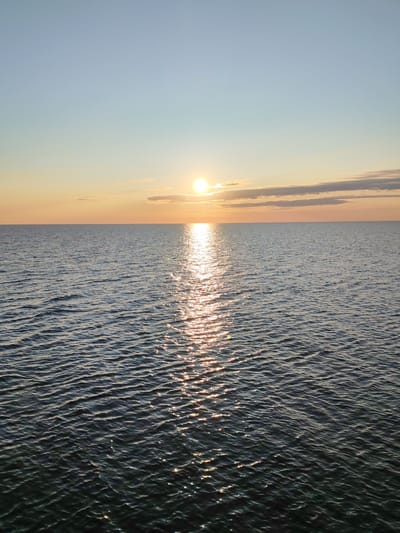 The image shows a tranquil body of water, likely a sea or large lake, under a clear sky with a faint haze near the horizon. The sun is setting, casting a warm, golden light that reflects off the gentle ripples on the water's surface. The sky transitions from a pale blue at the top to a soft orange and yellow near the horizon, indicating the time of day is late afternoon or early evening. There are a few wispy clouds scattered across the sky, also illuminated by the setting sun. The water is a dark, deep blue with subtle waves that catch the light, creating shimmering patterns. The horizon line is sharp and distinct, separating the sky from the water. The scene evokes a sense of peace and natural beauty. There are no people, buildings, or any signs of human activity visible in the image, suggesting a remote or natural setting. The image appears to be a landscape photograph taken outdoors.