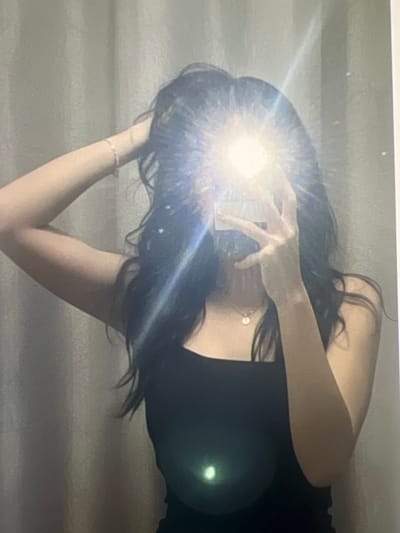 This image, taken in Ittenheim, France, captures a single individual performing a mirror self-portrait. The subject, exhibiting long, dark, wavy hair, is partially obscured by a bright photographic flash emanating from a handheld smartphone. The individual wears a black tank top, a gold-colored chain necklace with a circular pendant, and a thin gold-colored bracelet on the left wrist. The right hand holds the illuminating smartphone, while the left arm is raised with the hand near the hair. The background consists of a light-toned, vertically-folded curtain or textile. A distinct green lens flare is visible in the lower central area of the frame.