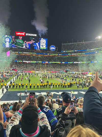 The image depicts a Super Bowl celebration at Levi's Stadium. The Jumbotron displays "SUPER BOWL LX CHAMPIONS" with images of players. Confetti rains down on the field, where players in yellow jackets stand. Spectators are seated in the foreground, many holding up phones to capture the moment. Someone wears a #31 Chancellor jersey. The crowd fills the stadium, extending upwards. A smokey haze is visible in the left portion of the image.