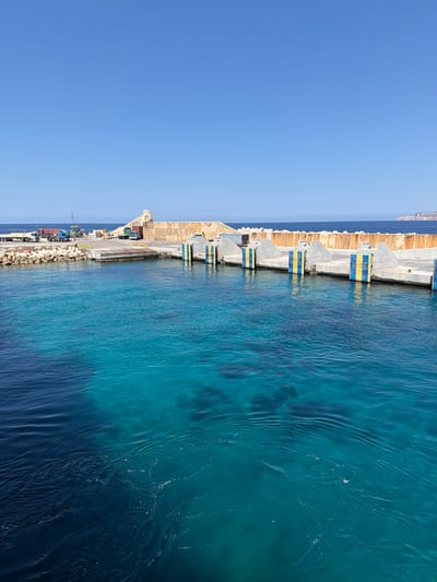 The image depicts a harbor scene in Mellieħa, Malta. In the foreground, the clear, turquoise waters of the Mediterranean Sea are visible, with darker blue water further out. The seabed is discernible in the shallower areas. A concrete pier extends into the water, lined with several mooring bollards painted with alternating yellow and blue stripes. Behind the pier, a large, textured concrete wall forms the main structure of the harbor. On the pier, several trucks and what appears to be a tractor are parked. Further back, the landscape rises with a rocky shore and a small, arched stone structure at the top of a wall. The sky above is a clear, cloudless blue. The scene suggests a functional port area with maritime activity.
