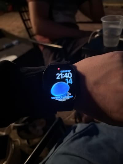 A close-up shot shows a person's wrist wearing a smartwatch displaying the time 21:40 and the date 14. The watch face also shows "CLEAR 21°" at the top, indicating clear weather and a temperature of 21 degrees. Below the time and date, a blue jellyfish graphic is visible, with a metric below it reading "136 BPM, 9 MIN AGO". The background is dark and out of focus, with hints of furniture and other people. The setting appears to be an indoor or outdoor area at night. The image does not provide any information about the location of Tankovo, Bulgaria.