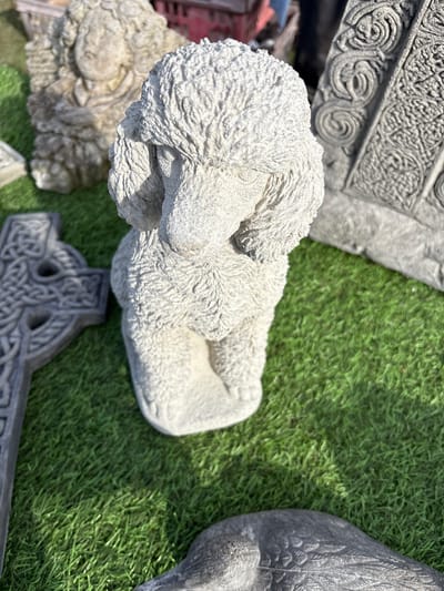 A high-angle shot captures a collection of ornamental stone objects situated on artificial turf. A reclining stone dog figurine, textured to mimic fur, dominates the center. Adjacent to it, a Celtic cross with intricate knotwork is visible. To the upper left, a stone slab with raised, swirling patterns is partially visible. An indistinct stone sculpture lies to the lower-left of the dog. To the right of the dog figurine is the body of a stone sculpture bird, with a grey color. The scene takes place on artificial turf, and it is a likely photograph of stone ornaments being sold in Chirk, United Kingdom.