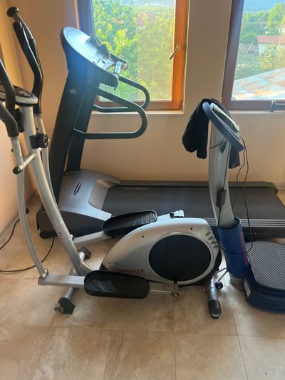 This media file shows a collection of exercise equipment, specifically an elliptical trainer and a treadmill, positioned indoors. The elliptical trainer is in the foreground, a silver and black machine with visible handles and foot pedals. The treadmill is behind it, a larger black machine with a textured running surface. Both machines appear to be in a room with tiled flooring. Natural light streams in from large windows on the left, illuminating the scene. Through the windows, trees and hints of rooftops are visible, suggesting a residential setting, possibly in Krainitsi, Bulgaria, as the prompt indicates. The lighting suggests it is daytime, and the overall atmosphere is calm and static, with no people present or any activity taking place. There is some text visible on the equipment, including "PRO-FORM" on the treadmill and "SPORTER" on the elliptical, along with other smaller labels and model numbers. The scene is a typical home or gym environment where fitness equipment is stored.