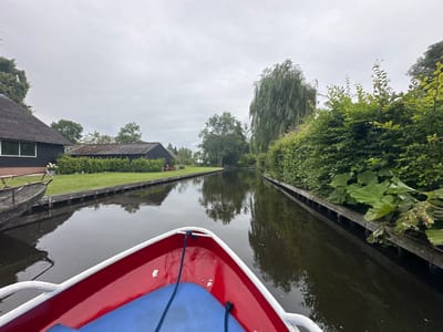 This is a POV shot from a red and white boat with a blue seat, moving down a narrow canal in Giethoorn, Netherlands. The canal is flanked by wooden docks and lush green foliage. On the left side of the canal, there are grass lawns and dark-colored buildings with thatched roofs. On the right side, the vegetation is dense, with large green leaves protruding over the wooden dock. The sky is overcast and gray. The water in the canal is dark and reflects the surrounding trees and buildings. There are no people visible in the frame. The scene depicts a tranquil, picturesque waterway characteristic of Giethoorn, a village known for its canals and lack of roads.