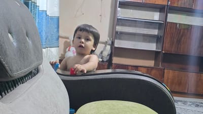A shirtless young boy is seated at a table, holding a red toy block in his right hand and a blue toy block in his left. He is looking towards the camera with a surprised expression. In the background, a stuffed animal resembling a kangaroo sits on a wooden shelf. To the left of the boy, a plush toy with a red heart is visible. The room is decorated with blue curtains. The overall environment suggests a domestic setting, likely in a city.
