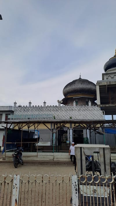 The image shows a mosque with a large, dark, dome-shaped roof. The mosque has white walls and several small spires. A sign in front of the mosque reads "MESJID BESAR PEUSANGAN". A man in a white shirt and dark pants stands to the right of the mosque, looking towards the camera. In the foreground, there is a fence made of white metal with circular designs. A motorcycle is parked near the mosque. The sky is overcast. The scene appears to be set in Pantegajah, Indonesia, a location hinted at by the sign on the mosque. The overall impression is of a quiet, possibly religious, scene in a developing area.
