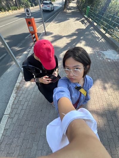 Two individuals are standing on a paved sidewalk next to a road in Ospedaletti, Italy. The person on the left wears a red baseball cap, a black t-shirt, and a hydration pack. They are looking down at their phone. The person on the right wears a blue button-up shirt, glasses, and a yellow strap across their chest. They are looking directly at the camera. In the background, a white SUV is driving on the road, and a speed camera is visible on the side of the road. Trees and a metal fence line the opposite side of the road. The ground is paved with hexagonal bricks, and shadows from trees and a railing are cast across the sidewalk. A portion of the photographer's arm and white clothing is in the foreground, obscuring part of the scene.