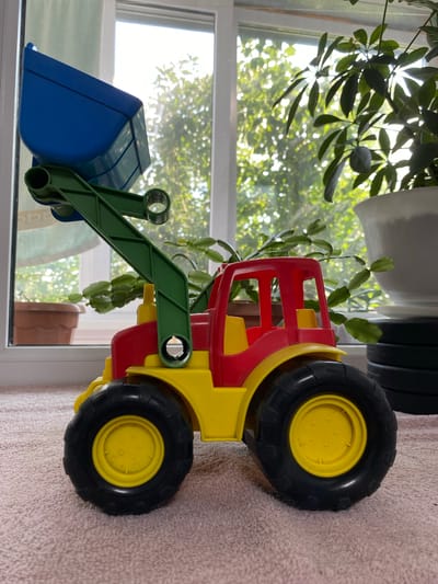 A bright red and yellow toy tractor with a blue bucket on a green arm sits on a pink carpet. The tractor has large black tires with yellow hubs. In the background, a window overlooks a sunny outdoor scene with green trees and foliage. There are also houseplants visible, one in a large white pot and another in a smaller brown pot. The scene is well-lit by natural light streaming through the window. There is no indication of the specific location in Бескөл ауылдық округі, Kazakhstan.