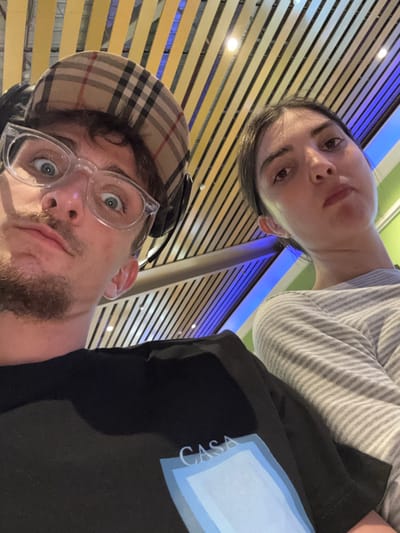 The image depicts two individuals in a low-angle, selfie-style shot, set indoors in Kasarvarne, India. In the foreground on the left, a male subject with short brown hair, facial stubble, and clear-framed glasses looks directly towards the camera with wide eyes and puckered lips. He is wearing a tan baseball cap with a dark plaid pattern, black over-ear headphones, and a black t-shirt featuring a light blue and white rectangular graphic with the word "CASA" printed in dark letters above it. To his right, partially obscured and slightly behind, a female subject with dark hair looks towards the camera with a neutral expression. She is wearing a light-colored long-sleeved shirt with horizontal stripes. The background reveals a modern indoor environment, characterized by a ceiling composed of parallel, light-colored slats with embedded circular light fixtures. Sections of blue accent lighting and a green panel are visible in the upper right background.