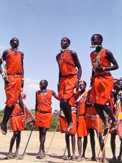 The image displays a group of approximately ten dark-skinned individuals, predominantly male, engaged in a synchronized vertical jumping dance in an outdoor environment in Ihumwa, Tanzania. Three central figures are suspended mid-air with knees bent, while others stand on the light brown ground. All individuals are attired in draped red garments, some cinched at the waist, and wear elaborate beaded necklaces, bracelets, and other adornments. Several standing individuals hold thin wooden staffs. The background features blurred green vegetation under a clear blue sky, indicating bright daylight conditions.