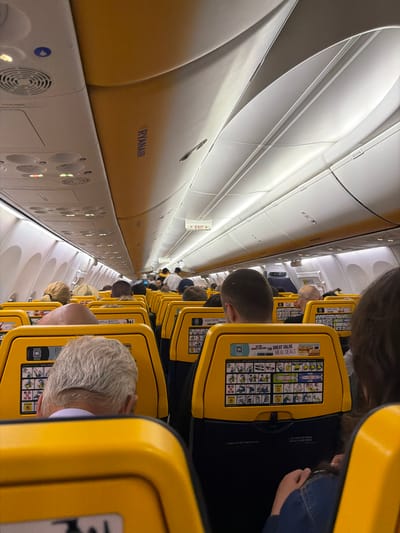The image shows the interior of a Ryanair airplane, likely in flight. The camera is positioned in the aisle, facing towards the front of the plane. Rows of yellow seats are visible on the right side of the frame, occupied by passengers with the backs of their heads in view. The overhead compartments are on the left, with a prominent "RYANAIR" logo. An exit sign is illuminated above the aisle. The image suggests a typical airplane cabin environment with passengers seated, during a Ryanair flight. There's no specific information in the image or its OCR to directly associate it with Luqa, Malta.
