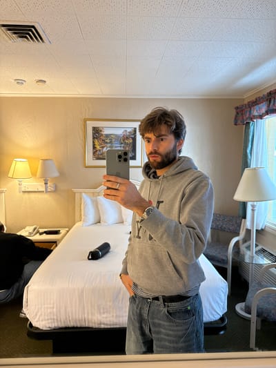 A person with brown hair and a beard is standing in what appears to be a hotel room, holding a gray smartphone to take a selfie in the mirror. The room contains a bed with white linens, pillows, and a black water bottle. There is a lamp beside the bed, artwork on the wall, and a window with a patterned curtain. A vent is visible on the wall, and an air conditioning unit is placed near the window. The person is wearing a gray hooded sweatshirt, a watch, and blue jeans. The location might be in a hotel room in Laconia, United States of America.