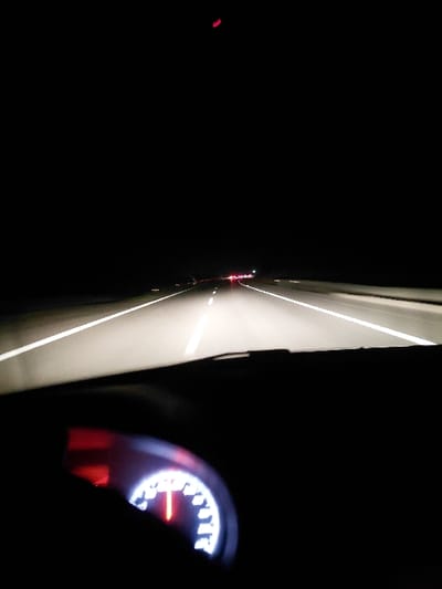 A nighttime view from inside a car shows the road ahead with glowing white lines. Tail lights of a distant vehicle are visible. The dashboard speedometer, illuminated with white numbers and a red needle, is partially obscured by a dark silhouette. The scene is predominantly dark.