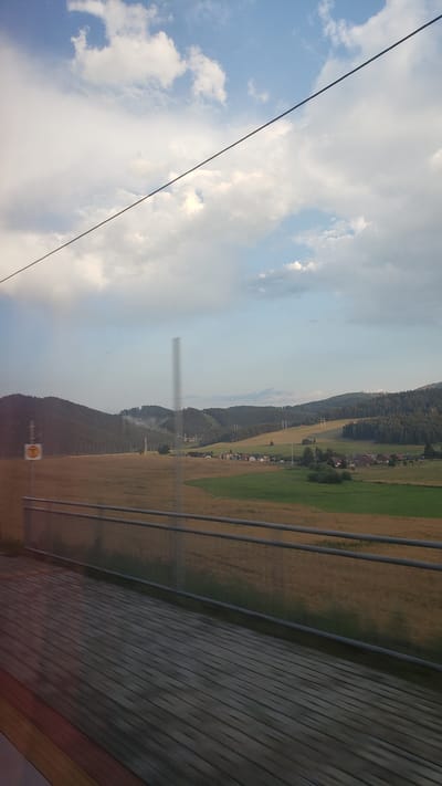 The image depicts a rural landscape in Lučivná, Slovakia, as seen from what appears to be a moving train. In the foreground, a wooden platform is visible, with a metal railing running along its edge. Beyond the railing, a vast field of golden-brown crops stretches towards a gently rolling hillside. The hillside is covered with a mix of trees and open, grassy areas, with some structures and power lines dotted across the landscape. In the middle ground, a small cluster of houses forms a village. The sky above is partly cloudy, with patches of blue sky peeking through. A thin wire stretches diagonally across the upper portion of the frame. The overall impression is one of serene natural beauty and agricultural activity in a Slovakian countryside setting.