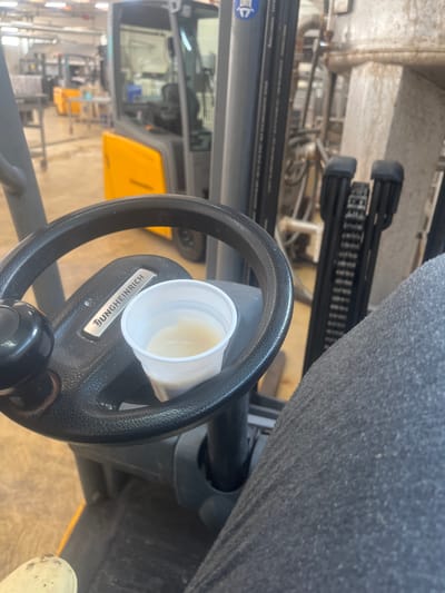 The image shows the interior of a forklift, with a white paper cup containing a light-colored liquid resting on the steering wheel. The steering wheel has a "Jungheinrich" logo. In the background, out of focus, a yellow and gray forklift is visible, along with industrial machinery and shelving. The environment appears to be a warehouse or factory setting. The image was taken from the perspective of someone seated in the forklift. There is no indication of the city of Cerignola, Italy in the image.