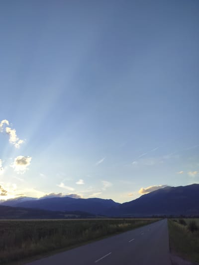 A paved road stretches into the distance, flanked by a field of tall, dry grass. In the background, a range of dark, silhouetted mountains is illuminated by the soft light of a setting sun. Wispy clouds are scattered across the clear blue sky, with dramatic rays of sunlight beaming through some of them. The scene is serene and natural, with no people or man-made structures visible besides the road itself. The time of day appears to be late afternoon or early evening. This image captures a rural landscape, likely on the outskirts of Manolovo, Bulgaria, given the mountainous terrain and open fields.