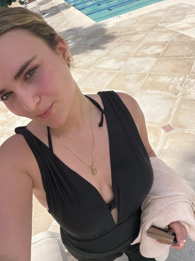A medium, eye-level, rotated, and extreme close-up shot captures a young woman with fair skin and blonde hair, seemingly at a poolside in Key Biscayne, Florida. The woman is wearing a black bathing suit or a dress-like garment with a revealing neckline and a gold necklace. She is holding a beige towel in her right hand, which also holds a black and bronze-colored device. In the background, a tiled pool area is visible. There is a chair with a white back support slightly behind the woman. The angle of the shot is canted, giving the impression that the woman is holding the camera above her and to the side.
