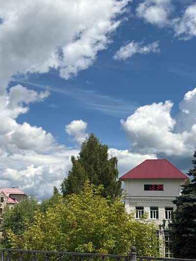This media file captures a bright, partly cloudy day in Pereslavl-Zalessky, Russia. The sky is a vibrant blue with large, fluffy white cumulus clouds interspersed with thinner, wispy cirrus clouds. The scene is dominated by a building with a prominent red-tiled roof. On the side of the building, a digital clock displays the time as 13:34, indicating it is mid-afternoon. Above the clock, a blue neon sign with Cyrillic lettering, likely an establishment name, is visible. Lush green trees, some with yellowing leaves suggesting late summer or early autumn, surround the building, partially obscuring it from view. A dark, ornate fence runs along the right side of the frame. In the distance, another building with a red roof is partially visible through the foliage. The overall impression is of a pleasant, sunny day with a hint of changing seasons.