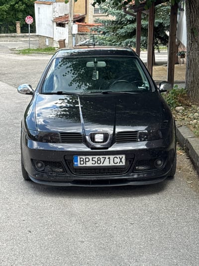 A black Seat Leon is parked on a paved street in Dobrinishte, Bulgaria. The car is facing the camera, with its front bumper and grille clearly visible. The license plate reads "BP 5871 CX". In the background, there are buildings, trees, and a stop sign. The sky is visible through the car's windshield, reflecting the trees and clouds. The scene appears to be a street in a residential area.