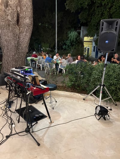 The image depicts an outdoor setting at night in Toritto, Italy, likely a restaurant or outdoor venue. A large tree trunk is visible on the left. In the foreground, there is musical equipment, including a keyboard on a stand, cables, and pedals. A large speaker on a tripod stands to the right, with a stage light on the ground below it. In the background, several people are seated at tables, engaged in conversation and eating. They are illuminated by ambient lighting. The environment appears to be a paved outdoor area surrounded by greenery and buildings.