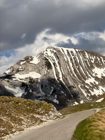 The image presents a view of a rocky mountain partially covered with snow, positioned alongside a narrow paved road. The mountain exhibits distinct horizontal layers, some accentuated with snow deposits. Patches of vegetation are sparsely scattered on the mountain's surface. Adjacent to the road, there's a slope covered with grass and rocks. A portion of the sky, filled with gray clouds, is visible in the background. The location is near the Boričje in Montenegro.