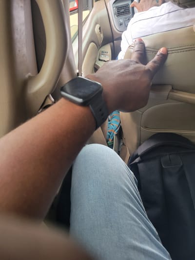 An African person wearing a watch on their left wrist is sitting in a vehicle, possibly a car or bus, in Katambe, Nigeria. Their left arm and hand are visible, with the hand making a peace sign. The person is wearing blue jeans. In the background, another person wearing a white and black striped shirt is visible, sitting in the same vehicle. A black backpack is on the seat behind the person's legs. The interior of the vehicle is visible, including beige upholstery and a car's air conditioning vent. The lighting suggests it is daytime.