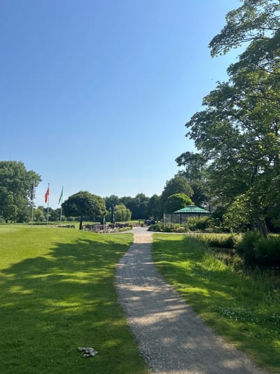 A gravel path leads through a lush green landscape, characteristic of a golf course in Wassenaar, Netherlands. The path curves gently to the right, bordered by manicured grass on the left and overgrown vegetation and a small body of water on the right. The sky above is a clear, bright blue, with the sun casting dappled shadows from nearby trees onto the path and grass.
In the distance, on the left side of the image, there are three flags on poles, two of which are distinctly colored red and green, suggesting a national or club affiliation. Beyond the flags, a treeline forms the horizon, with some buildings or structures partially visible amongst the foliage. Closer to the path, on the left, several sets of outdoor tables and chairs are arranged under umbrellas, some of which are green, suggesting a seating area or patio associated with the golf course.
On the right side of the image, further back from the path, a distinctive green umbrella with a logo resembling "Heineken" is visible, suggesting an outdoor bar or seating area. Several golf carts are parked near this area, indicating active use of the golf course. The overall impression is of a serene, sunny day at a well-maintained recreational facility.