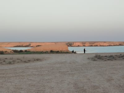 The image depicts a serene natural landscape in Haratan, Uzbekistan, at dusk. In the foreground, a vast expanse of dry, sandy terrain stretches across the frame, sparsely dotted with small clumps of dry vegetation. A dirt track, possibly an unpaved road, winds its way through the sand towards the middle ground. In the mid-ground, a calm body of water, likely a lake or a calm section of a larger body of water, meets a sandy shore. Beyond the water, a series of low, rounded hills with reddish-brown rocky surfaces form the horizon. The sky above is a gradient of pale colors, transitioning from a light blue near the horizon to a soft, almost white hue at the top of the frame, indicating the twilight hours.
Several individuals are present in the scene. On the far right, a lone person wearing dark clothing stands near the water's edge, facing away from the camera. Closer to the center, three figures are visible near the shoreline. One individual, also in dark clothing, appears to be walking or standing. To their left, two other figures are seated or crouching on the sand. The overall impression is one of quiet contemplation and natural beauty at the close of day in this Uzbek location.