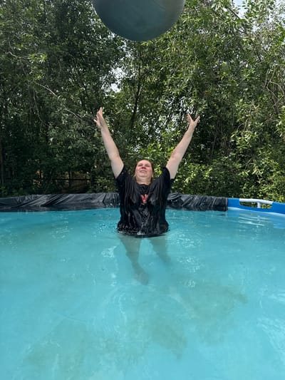 A person is standing in a swimming pool, with their arms raised. The person is wearing a black t-shirt, which appears to be wet. The pool is filled with blue water, and the bottom of the pool is visible. The person is positioned in the center of the frame, and their body is partially submerged in the water. Behind the pool, there are trees and a fence. The sky is visible above the trees. A large, grey, oval-shaped object, possibly a large ball, is partially visible at the top of the image. The scene appears to be outdoors, possibly in a backyard. The location is identified as the city of Хохряки, Russia.