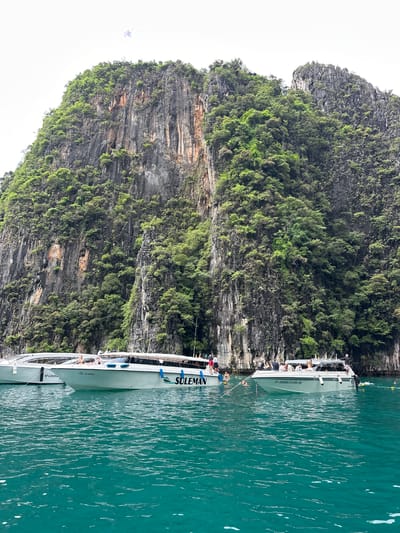 A scenic view of towering, lush green cliffs rising from a turquoise sea. Two white speedboats are anchored near the shore, with several people visible on board and in the water. The boat on the right has "SULEMAN" written on its side. The cliff face is rugged and covered in vegetation, with some exposed rock. The water is clear and calm, with gentle ripples on the surface. This scene appears to be from a tropical location, possibly Haad Yao, Thailand, known for its limestone karsts and clear waters.