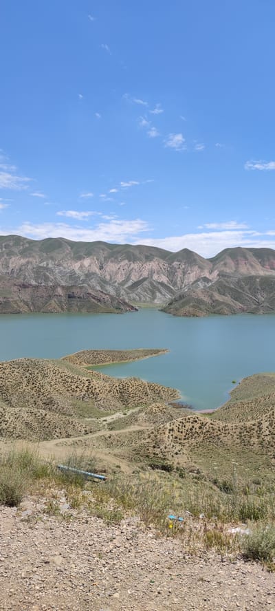 The image depicts a landscape with a lake and mountains under a partly cloudy blue sky. The foreground shows a rocky, arid terrain with sparse vegetation. A blue pipe lies partially hidden among the foliage and rocks. The midground features a lake with a greenish-blue hue, surrounded by hilly terrain covered in dry, scrubby vegetation. A peninsula extends into the lake. In the background, rolling mountains are visible, with varied coloration including green, brown, and reddish tones. The landscape suggests a natural setting in a region with an arid climate. This is Lanjazat, Armenia.