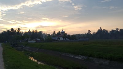 The image depicts a rural landscape in Blangtingkeum, Indonesia, at sunrise or sunset. A dirt road runs along the left side of the frame, bordering a concrete irrigation canal. A grassy field stretches towards the center of the image. In the background, there are trees, a small house with smoke rising from it, and a white building with a dome, possibly a mosque. The sky is filled with clouds illuminated by the sun.