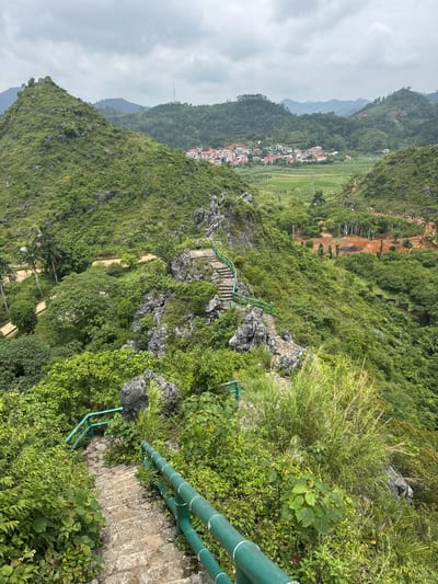 The image depicts a lush, mountainous landscape in Tam Thanh Ward, Vietnam. A stone staircase, bordered by a vibrant green railing, winds its way down a steep, vegetated slope. The stairs are made of irregular stone blocks and appear well-worn. Dense green foliage, including trees, bushes, and tall grasses, surrounds the path. In the distance, a cluster of red-roofed houses nestled in a valley is visible, surrounded by green fields and more rolling hills. The sky is overcast with grey clouds. The scene appears to be a scenic viewpoint or hiking trail within a natural park or rural area. There are no people visible in the image.