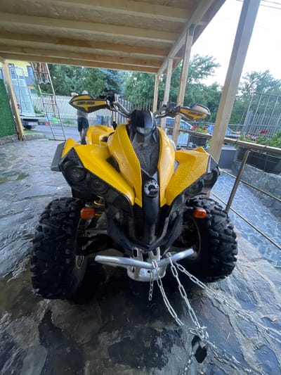 A yellow Can-Am ATV is parked on a wet stone patio under a wooden awning in Ivanyane, Bulgaria. The ATV is covered in water droplets. Two metal chains are attached to the front bumper of the ATV and are lying on the patio. In the background, a person is standing on a ladder next to a white fence and a building. Trees and other vegetation are visible behind the fence.