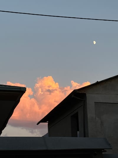 In Khujakent, Uzbekistan, at dusk, a serene scene unfolds. The sky, a canvas of soft blue and hues of pale orange, is punctuated by the ethereal glow of a crescent moon. The light catches the edges of voluminous clouds, painting them with vibrant peach and rose tones. Silhouetted against this celestial spectacle are the angular forms of rooftops and the stark lines of a utility wire stretching across the upper expanse. The architecture, characterized by its muted gray tones and sharp angles, provides a grounding contrast to the dynamic sky, suggesting a quiet moment of transition from day to night in this urban setting.