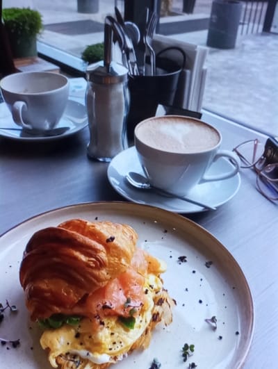 The image depicts a close-up, elevated view of a table setting within a cafe or restaurant in Yangiabad, Uzbekistan. In the foreground, a croissant sandwich is presented on a beige-rimmed white plate. The golden-brown croissant is sliced open and filled with scrambled eggs, several thin slices of orange smoked salmon, and green garnishes. The plate also shows scattered dark specks, likely seasoning, and small sprigs of herbs.
On the light-colored table, positioned to the right of the plate, is a white ceramic cup on a saucer containing a latte or cappuccino, identifiable by the heart-shaped latte art in its foam. A silver spoon rests on the saucer. To the left of this cup, a silver-capped glass sugar dispenser filled with white sugar stands next to an empty white ceramic cup on a saucer with a spoon. Further back, a black metal caddy holds an assortment of silver cutlery (forks, knives, spoons) and a stack of white paper napkins. A pair of eyeglasses with pink and gold frames rests on the table near the full coffee cup.
Through a large window in the background, an outdoor paved area is visible, featuring blurred green foliage in a planter, and indistinct structures including what appears to be a dark trash receptacle. The outdoor light suggests daylight conditions. No individuals are present in the scene.