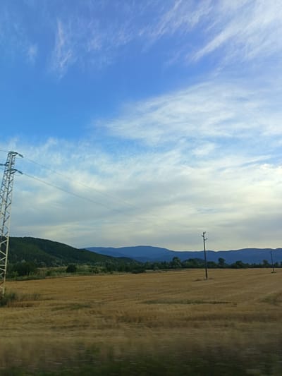 A wide shot captures a rural landscape with a vast, golden-brown field in the foreground. The field appears to be recently harvested wheat or barley, with stubble visible. In the middle ground, a line of dark green trees and a rolling, forested hill stretch across the frame. Beyond the hill, a range of blue mountains fades into the distance. A tall, metallic power line tower stands to the left of the frame, with wires extending across the scene. A single wooden utility pole is visible in the middle right, and another in the far right. The sky is a bright blue, filled with wispy white clouds that are streaked across the upper portion of the image. The overall impression is one of a serene and expansive countryside. The image was taken in Novachene, Bulgaria.