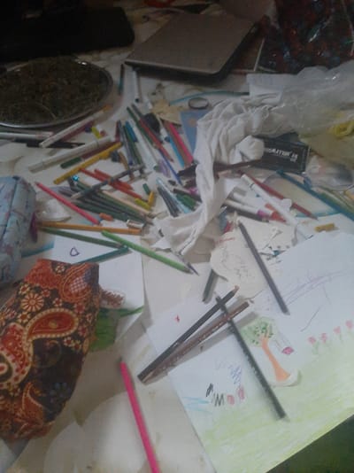 The image depicts a cluttered surface, possibly a table or floor, covered with various art supplies and personal items. In the center of the image, a pile of white cloth is spread out. Scattered around are numerous colored pencils, pens, and markers, some of which appear to be in use or have been used. There are also several sheets of paper with children's drawings. One drawing features a tree with a pink flower and a bird. Another appears to have scribbled text and shapes. A dark-colored tray filled with what looks like dried herbs or plant material is visible on the left side of the image. A dark laptop or similar electronic device is also present, partially obscured. A patterned fabric pouch, possibly a pencil case, is situated in the lower left corner. The overall impression is one of a creative workspace, perhaps for a child. The location is identified as Sabunchu, Azerbaijan, although no specific landmarks or context to confirm this are visible within the image itself.