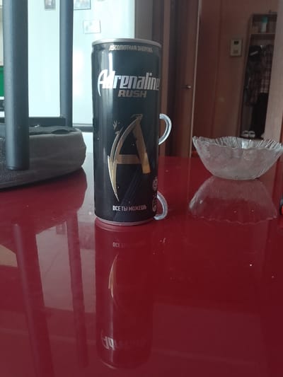 The image shows a close-up of an Adrenaline Rush energy drink can placed on a red, reflective surface, likely a table. The can is black with silver and gold lettering. The text on the can reads "АБСОЛЮТНАЯ ЭНЕРГИЯ" (Absolute Energy) and "Adrenaline RUSH". Below the logo, there's smaller text that says "ВСЕ ТЫ МОЖЕШЬ" (You Can Do Anything). The can has two plastic tabs attached to it. To the right of the can, there's a clear, decorative glass bowl with a wavy edge, resting on an inverted glass bowl or a similar object, creating a reflective effect. The background is out of focus but appears to be an indoor setting, with vertical structures that might be part of furniture or a room divider, and some pictures or frames on the wall. There are no people visible in the image. The lighting suggests it was taken indoors, possibly with natural light coming from the left. The overall scene is a still life composition featuring the energy drink and glassware. The specific location in Otradnaya, Russia, cannot be determined from the visual information in the image.