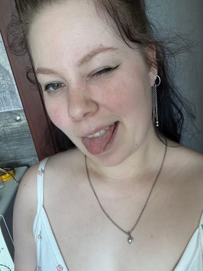 The image shows a close-up of a young woman winking and sticking out her tongue. She has fair skin with freckles, light brown hair, and is wearing a white tank top with a floral pattern, a silver chain necklace with a heart-shaped pendant, and a silver earring with dangling chains. Her left eye is open and she is wearing black eyeliner. The background is out of focus but appears to be an indoor setting with a wooden surface and a wall. There is a blurred object on the left that appears to be a container with a yellow lid. No specific location in Выльгорт, Russia is identifiable.