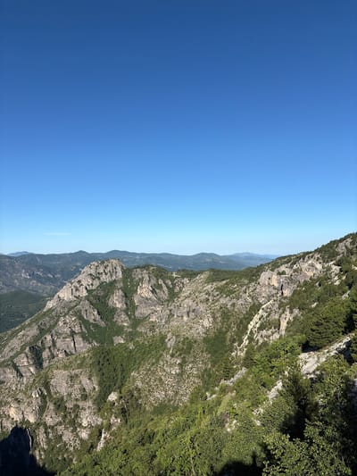 The image depicts a panoramic view of a mountainous landscape under a clear, bright blue sky. The foreground is dominated by a steep, rocky mountainside covered in dense green vegetation, including trees and shrubs. The rocks are textured and appear to be a light grey or beige color. In the mid-ground, more rugged mountains rise, also sparsely vegetated with patches of green. In the distance, a series of rolling hills recede, their outlines softened by atmospheric perspective, and they appear to be covered in a darker green foliage. The overall scene is one of natural, untamed wilderness. The location is identified as Kućišta, Montenegro. There are no people, animals, or man-made structures visible in the image.